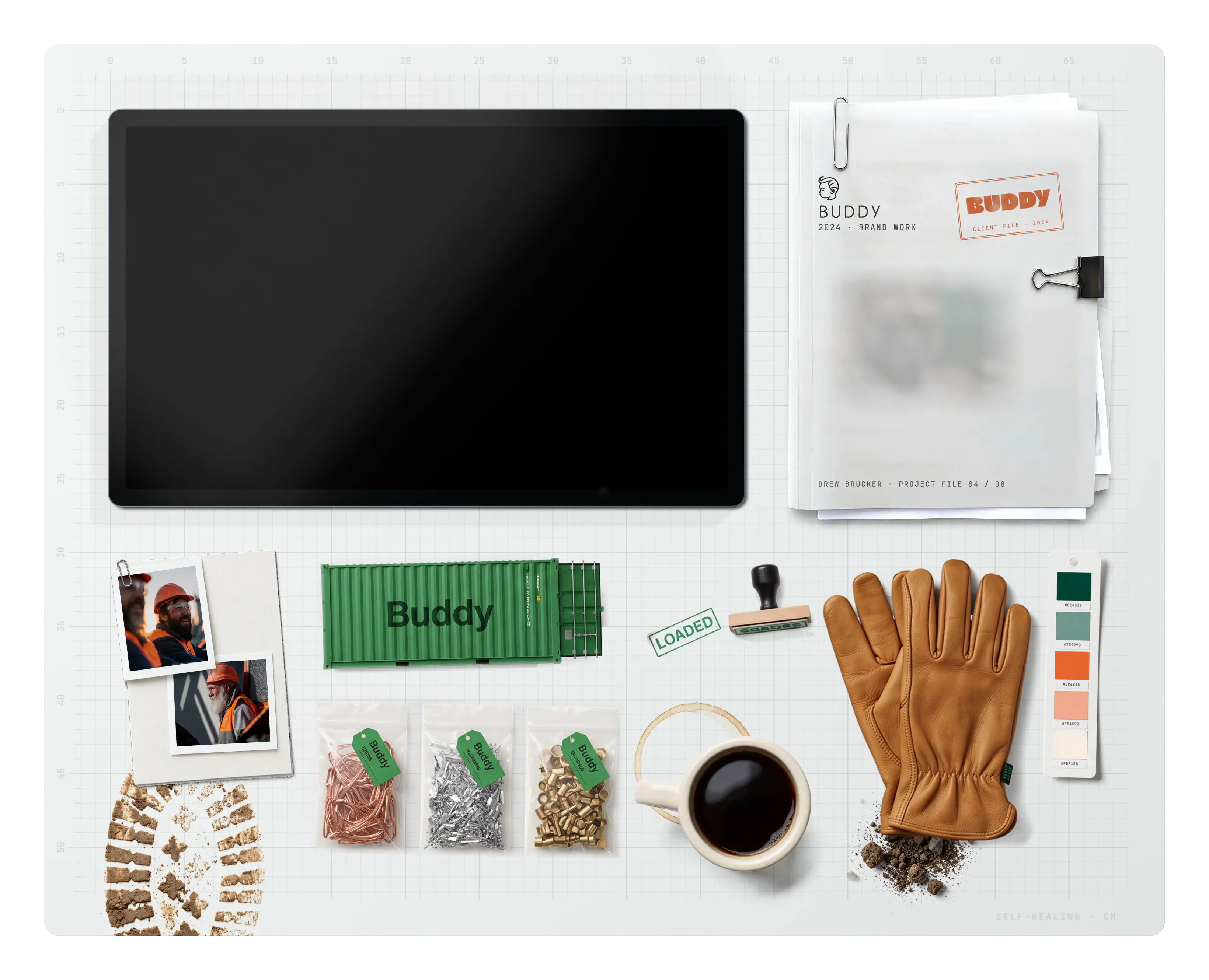 Buddy on the desk: the project file, a muddy boot print, clipped prints, a Buddy shipping container model, bagged scrap metal samples, a coffee ring, a mug of coffee, a LOADED stamp mark, a LOADED rubber stamp, a little dirt, leather work gloves and the brand swatch