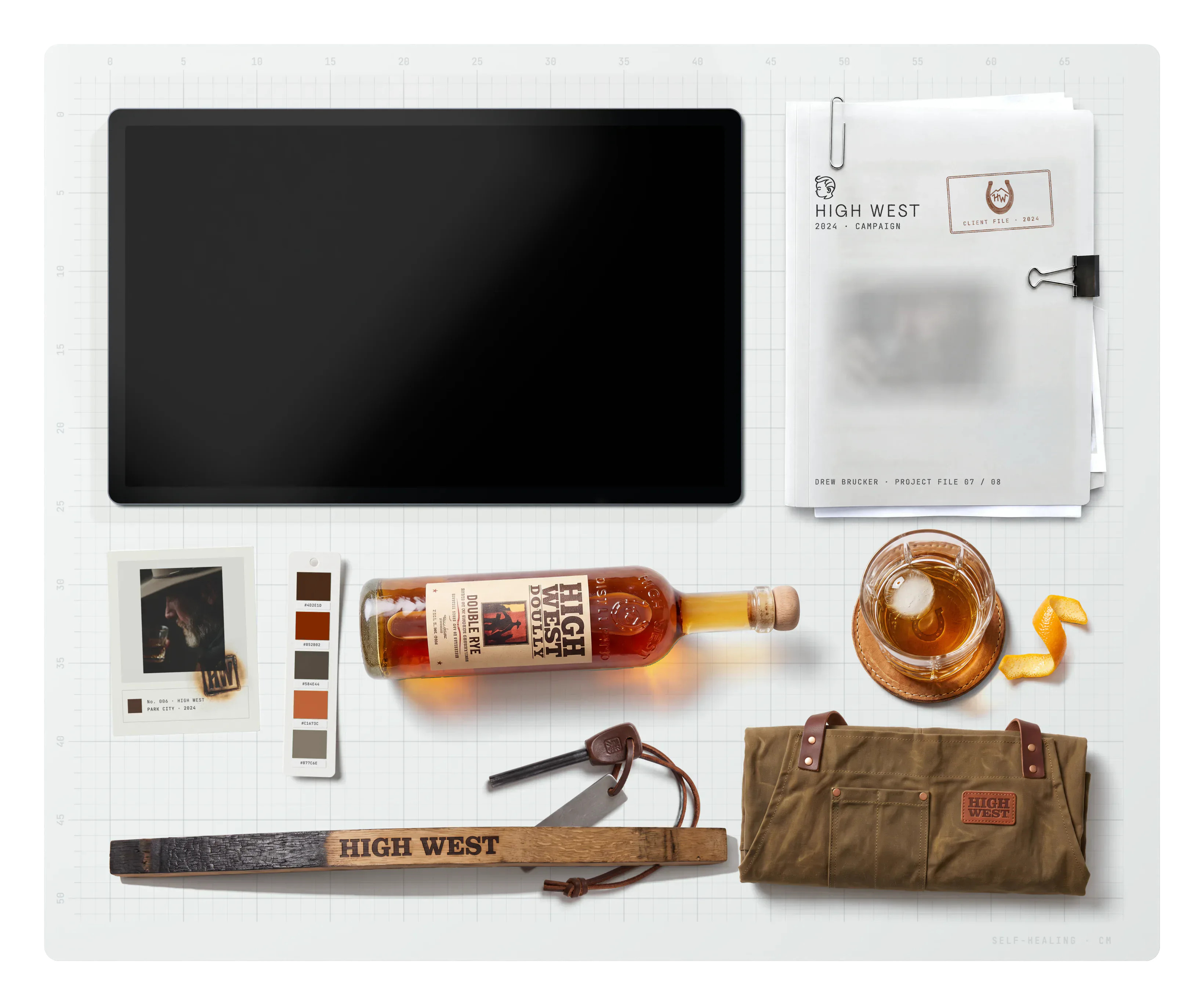 High West on the desk: the project file, archive cards, the brand swatch, a bottle of High West Double Rye, a whiskey on the rocks, an orange peel twist, a ferro rod fire starter, a charred barrel stave branded HIGH WEST and a waxed canvas apron roll